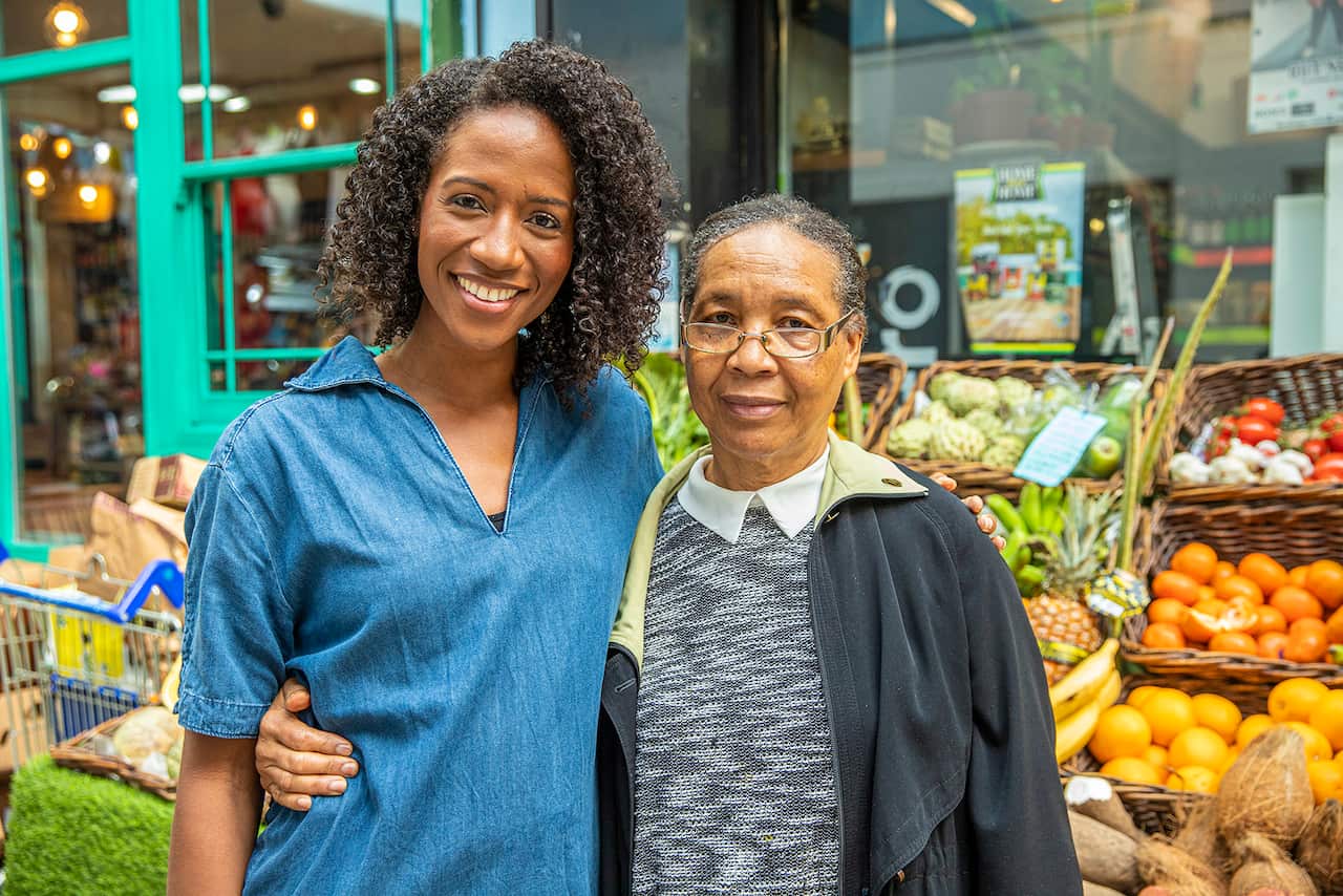 Two women stand in front of a fruit and vegetable stall. 