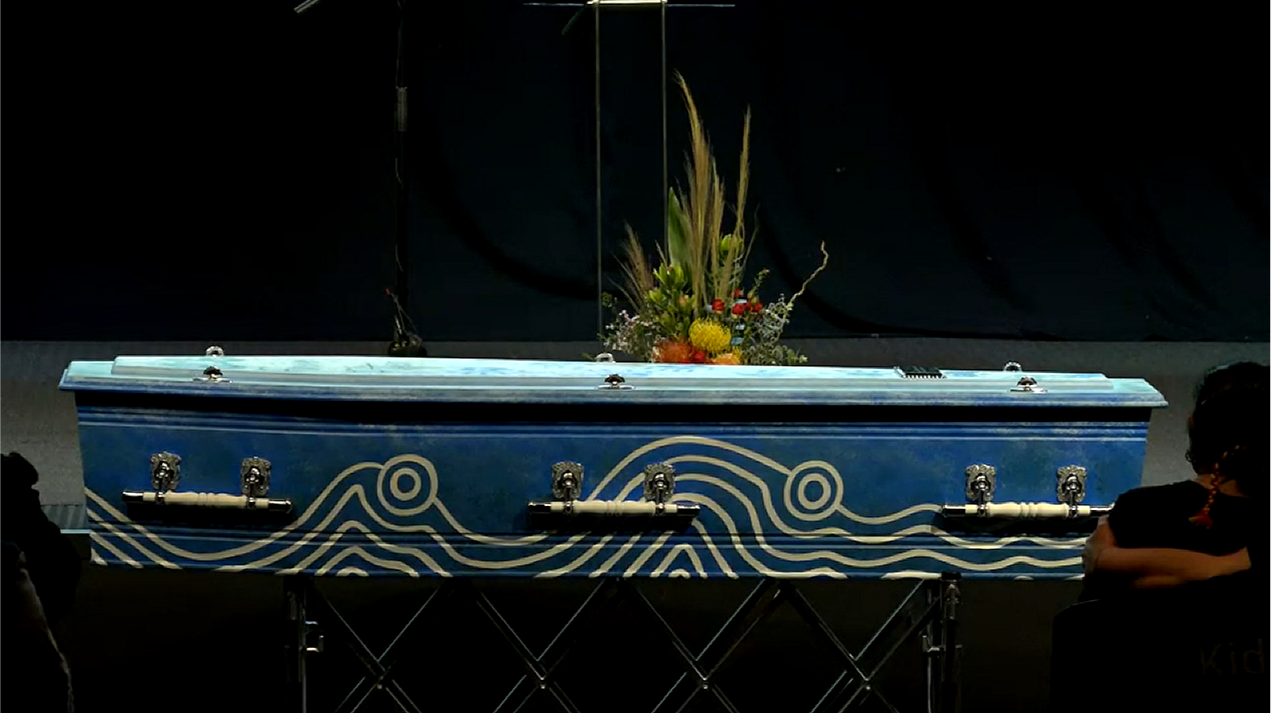 A small blue coffin, painted in white with traditional markings, sits on top of a gurney, with a small floral arrangement of natives sitting atop. 