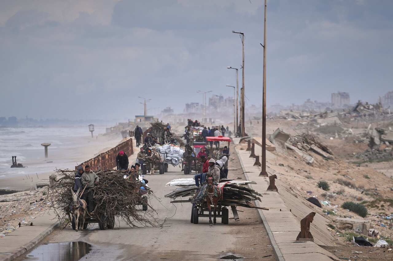 Several horse-drawn carts ferry people, their belongings, wood and other items along a beachside road.
