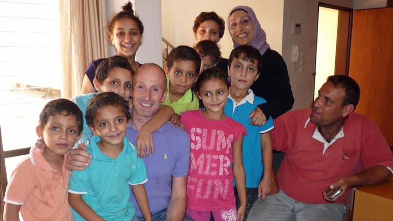 Seven pre-teen boys and girls wearing colourful t-shirts are huddling around a bald man in a purple polo shirt. Behind them stand two teenage girls and a woman in a hijab. On the side, a man in a red polo shirt is seated, looking at the family