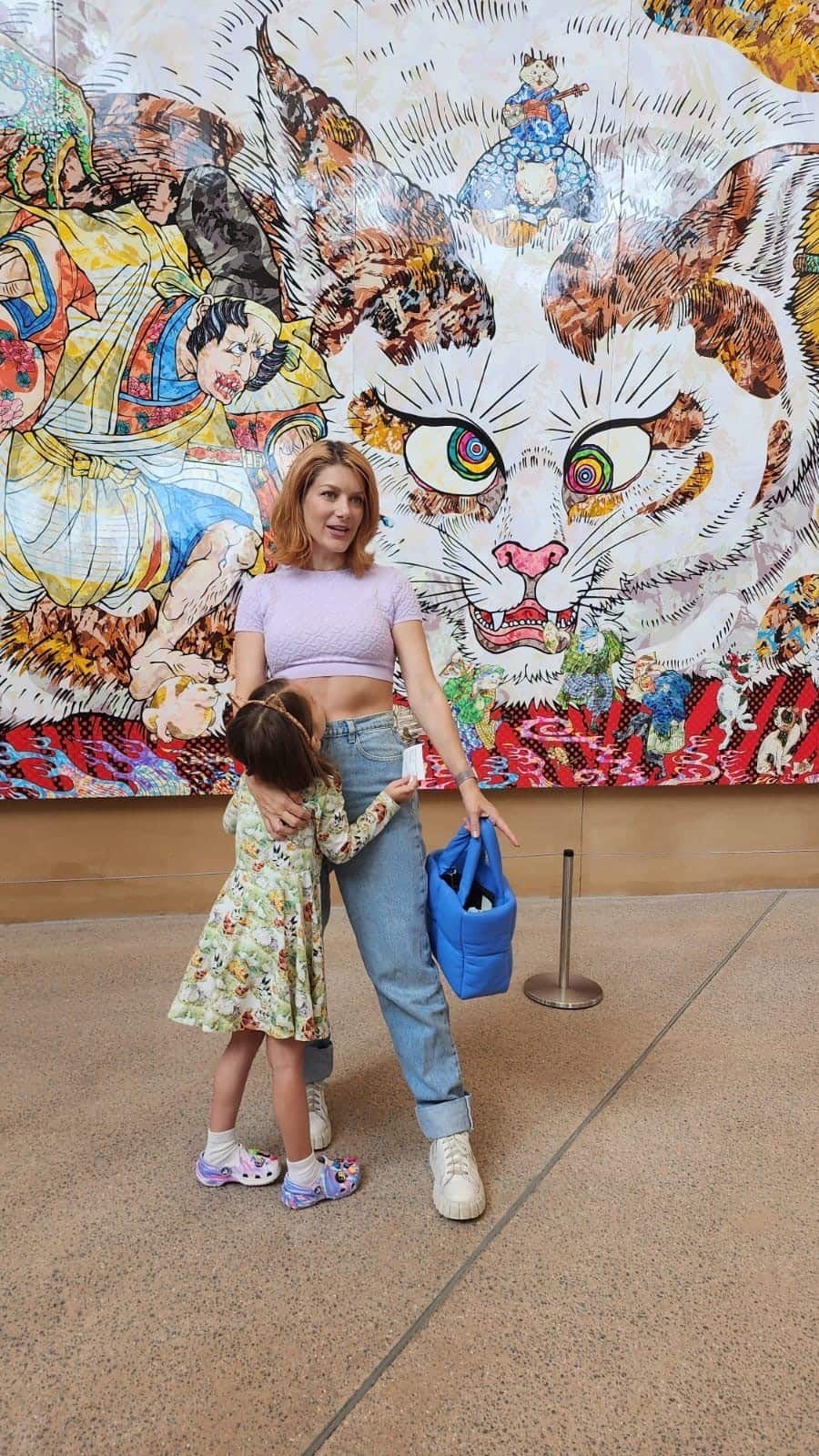 A woman dressed in jeans embraces a young girl while standing in front of a large painting outside.