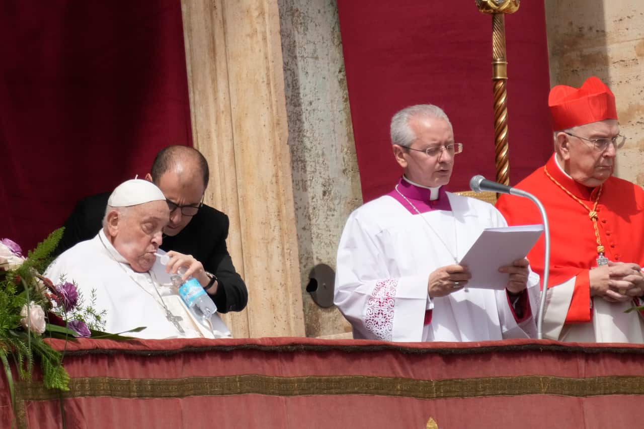 Pope Francis sitting down on a chair sips water next to a clergyman speaking into a lectern microphone.