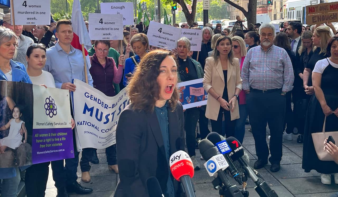 A woman in a blue suit speaks into microphones. Behind her is a crowd holding posters.