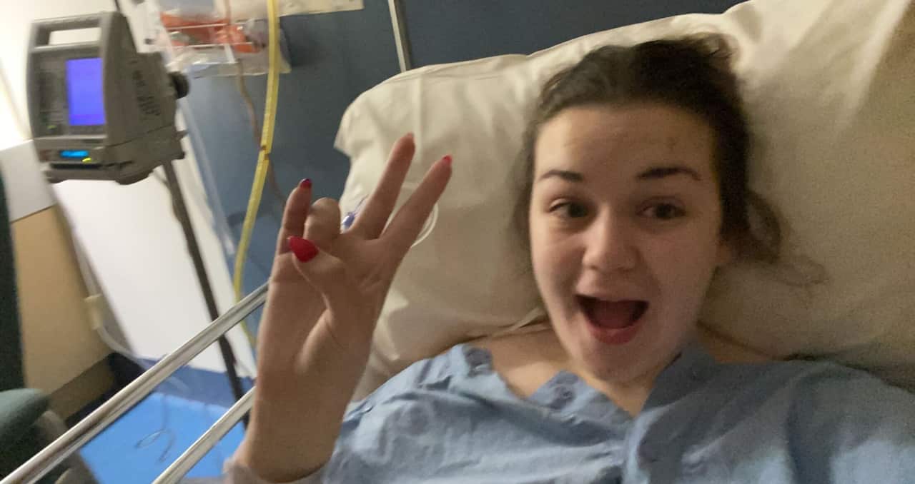 A brown-haired girl poses from a hospital bed, holding the peace sign aloft and smiling.