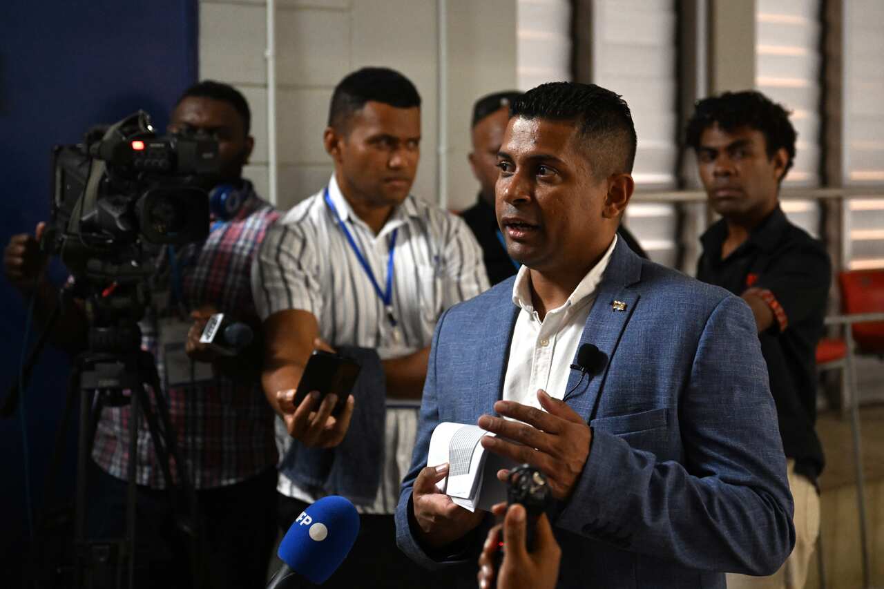 A man wearing a suit speaks at a press conference in front of cameras. 