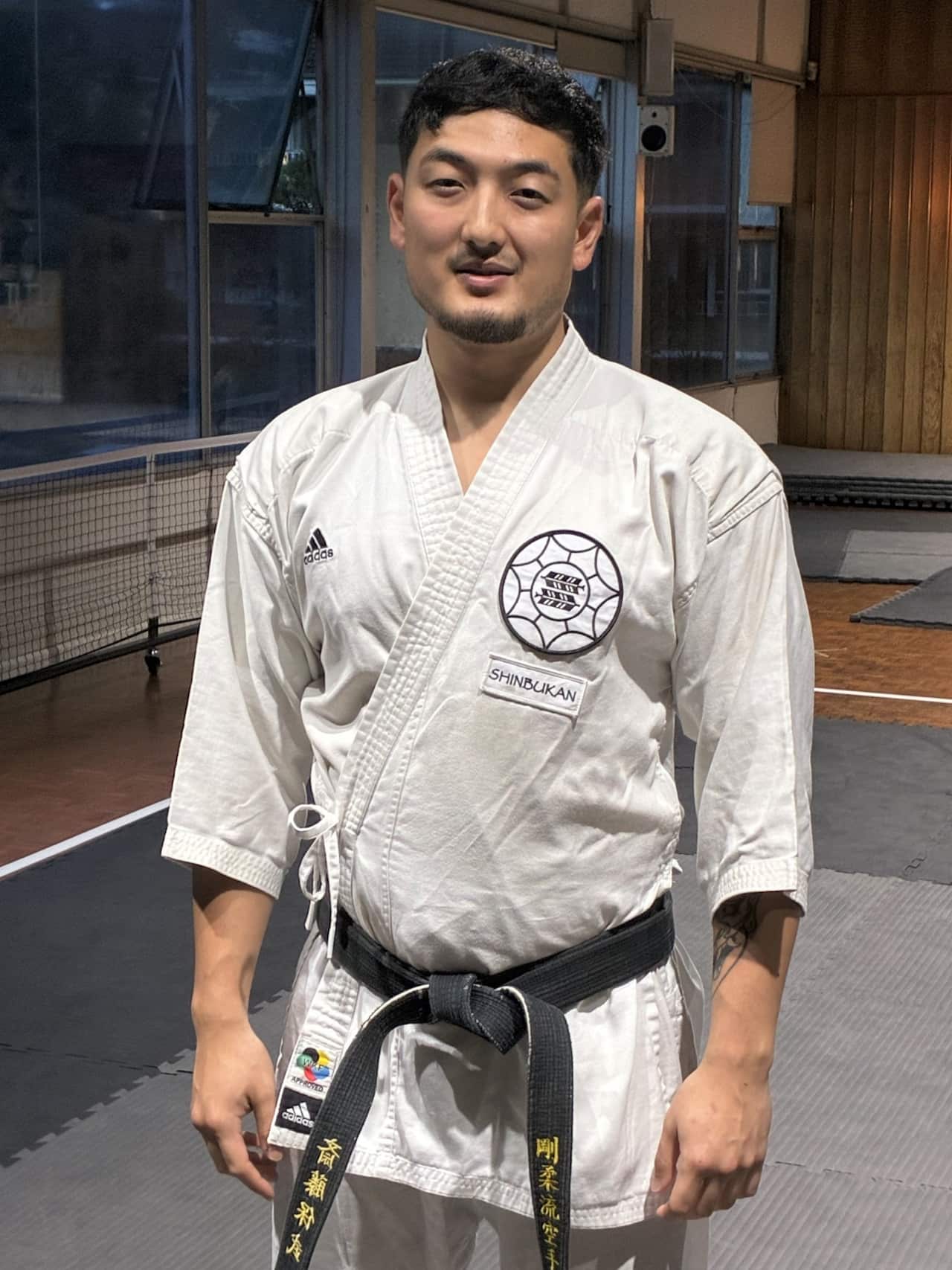 A man with black hair and a beard steands wearing a white karate jacket tied with a black belt.