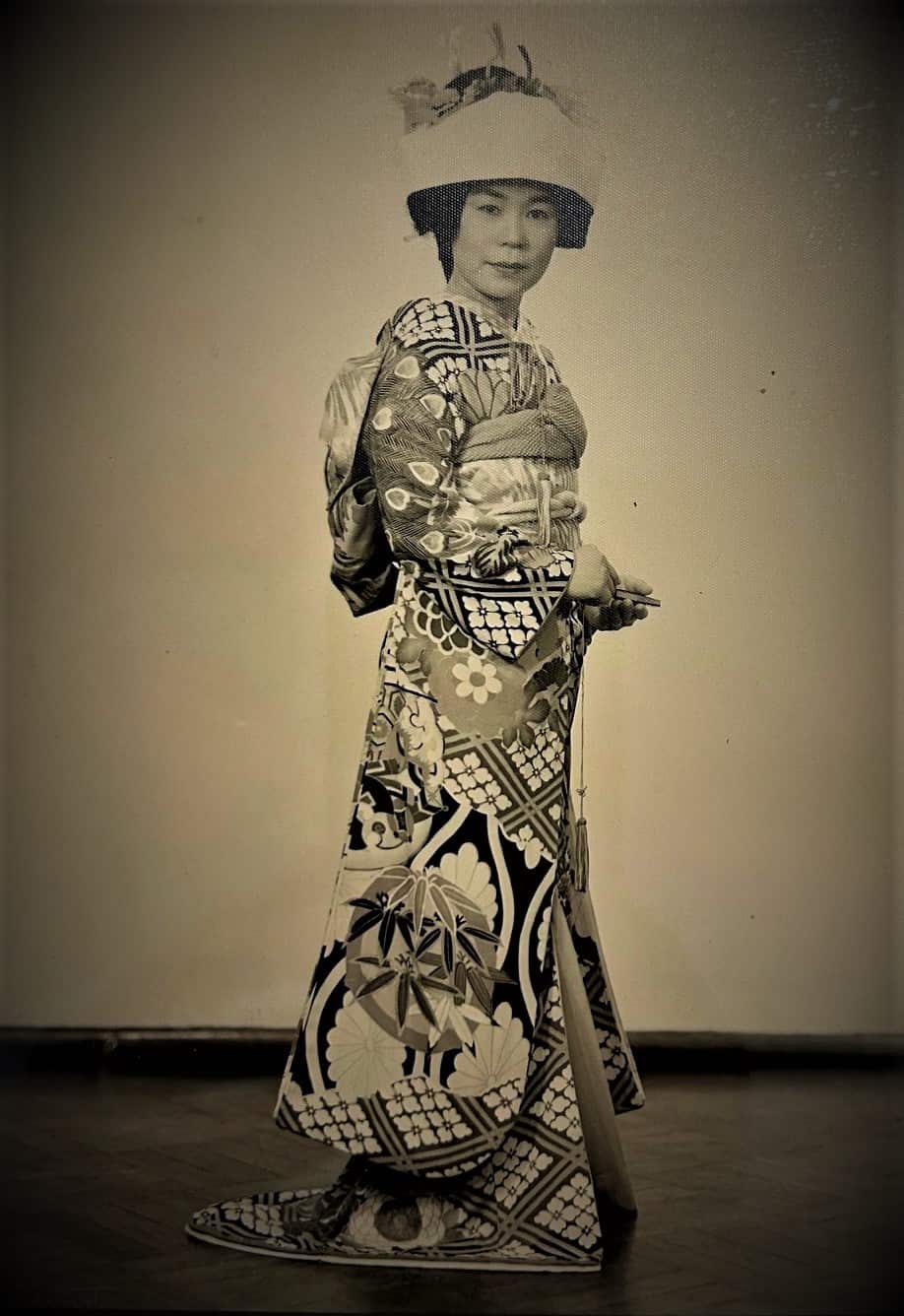 A woman in an elaborate kimono and hat stands facing the camera.