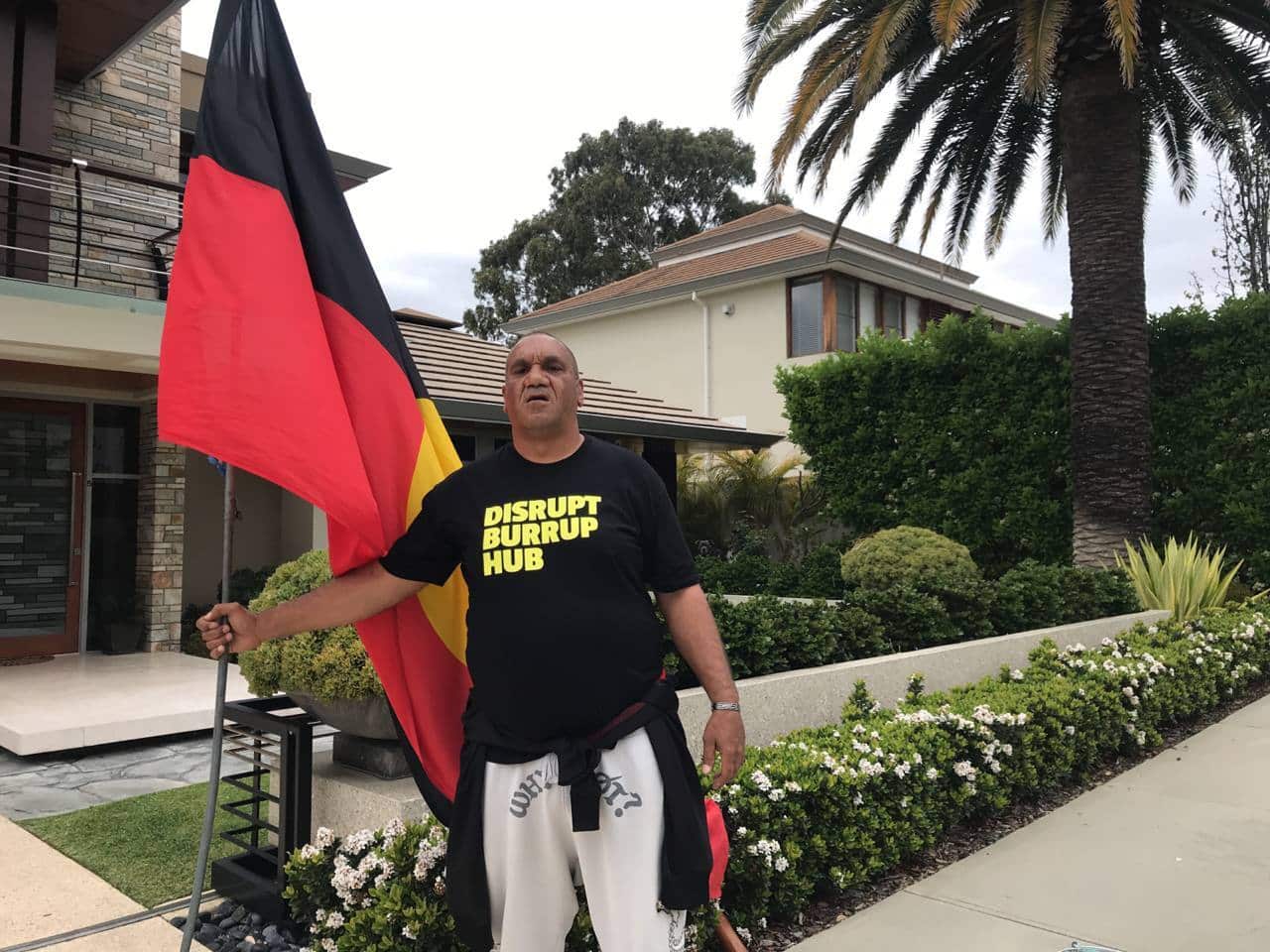 A man stands in front of a house, holding the Aboriginal flag. 
