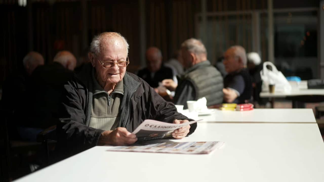 An old man in his 90ssitting at a table reading a newspaper. He is wearing a black outer jacket and an olive coloured inner jumper. Out of focus behind him are a dozen other old men sitting around.