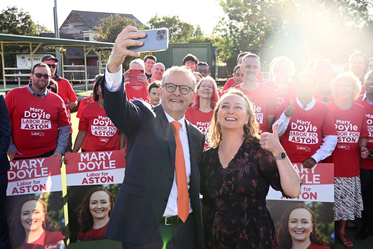 Man in a suit and glasses takes a selfie with a women giving a thumbs up sign.