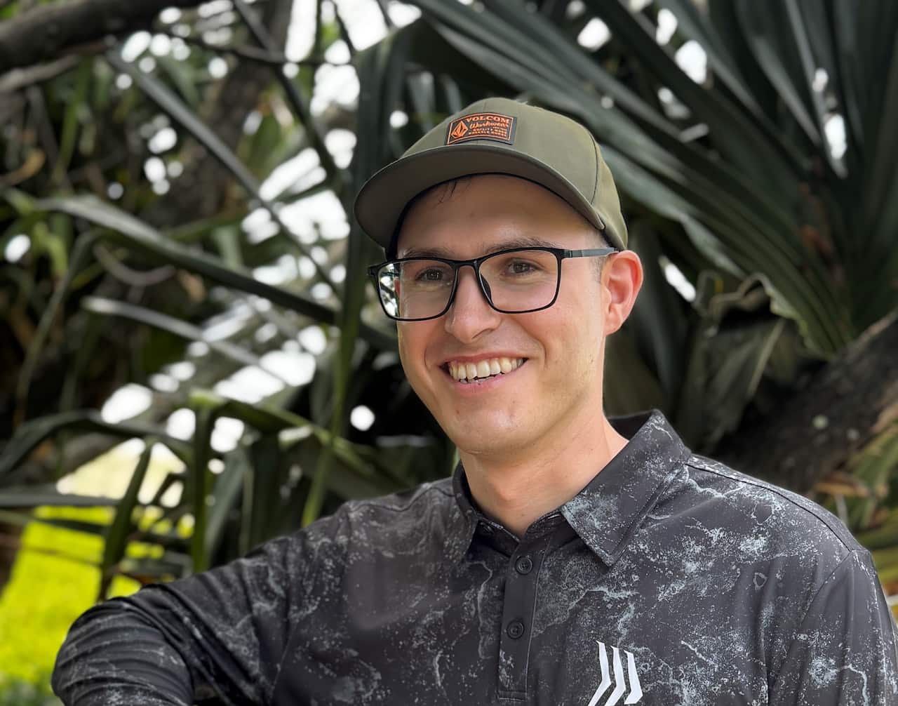 A man with glasses and a hat smiling outside.