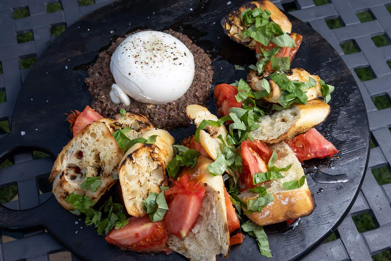 A round blcak tray holds a semiciclue of grilled bread and tomato, garnished with basil, sitting around a white ball of cheese on a round bed of olive dip. 