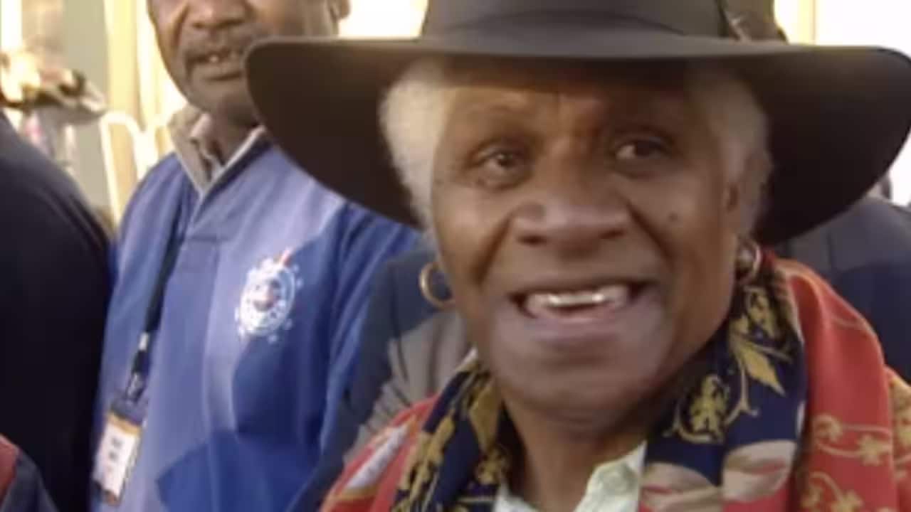 An elderly woman, wearing a hat and a colourful scarf around her neck, smiles.