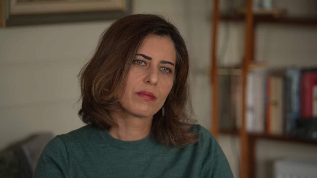 A woman with shoulder-length brown hair sits indoors on a couch, looking slightly off to the side with a serious, reflective expression. She is wearing a green sweater, and warm indoor lighting softly illuminates her face against a plain background.