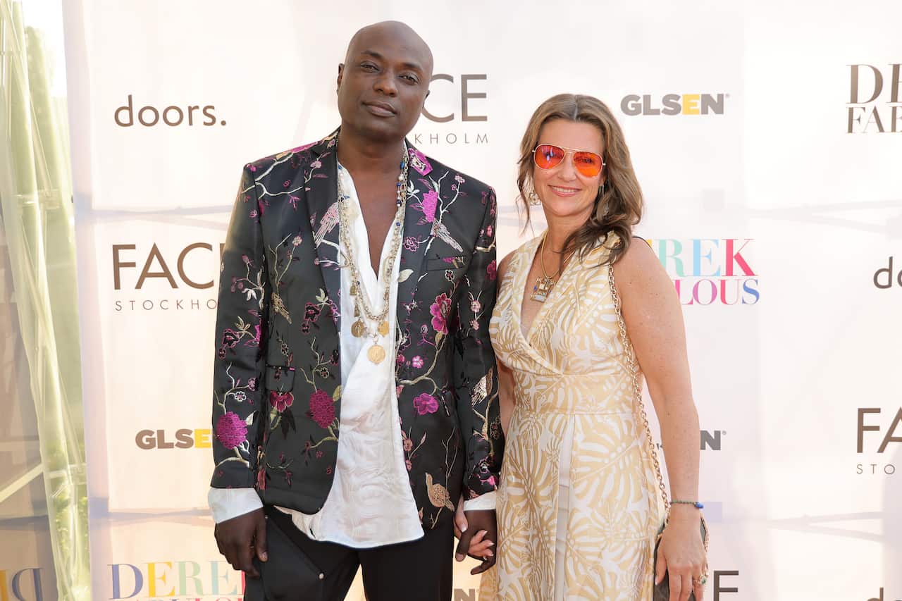 Man in floral blazer holds hands with woman wearing sunglasses and cream dress. 