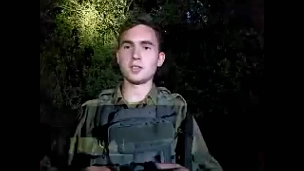 A young man in camouflage military gear standing somewhere dark, with a light cast directly on him.