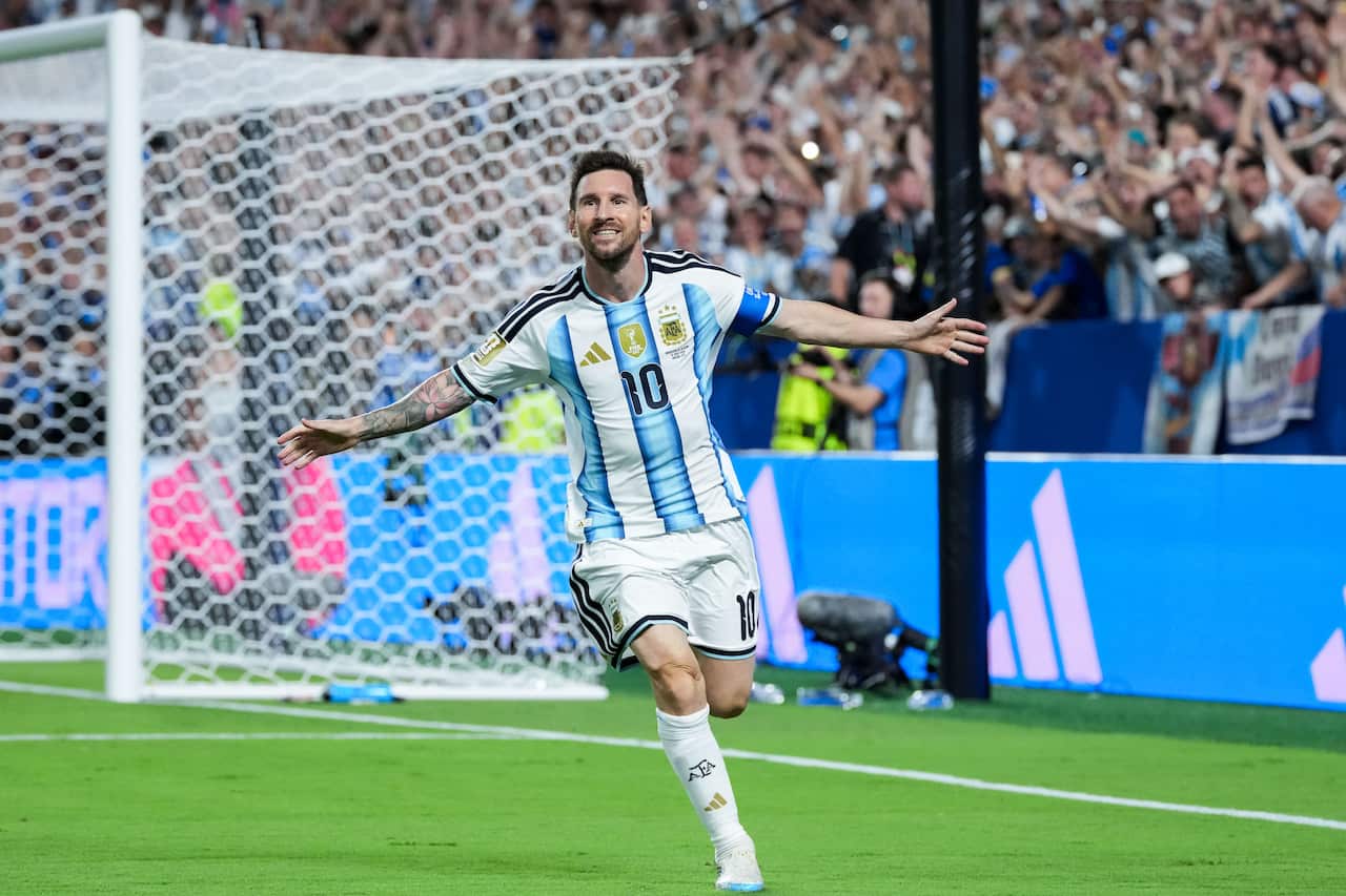 Lionel Messi, wearing a light blue and white jersey, runs away from the goal with his arms outstretched