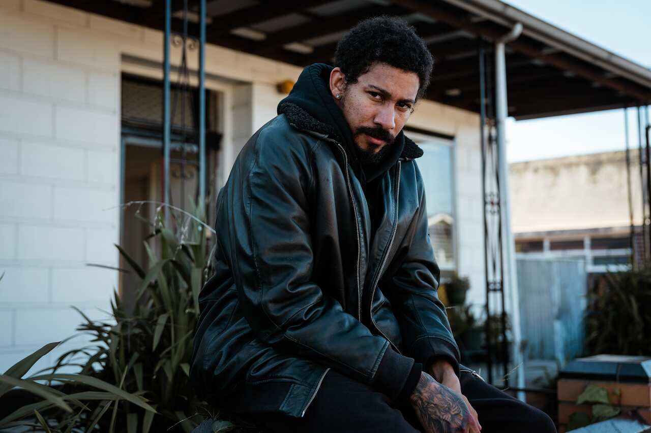 A man with a moustache, in a leather-look jacket, sits outside a house.  A tattoo can bee seen on one hand. 