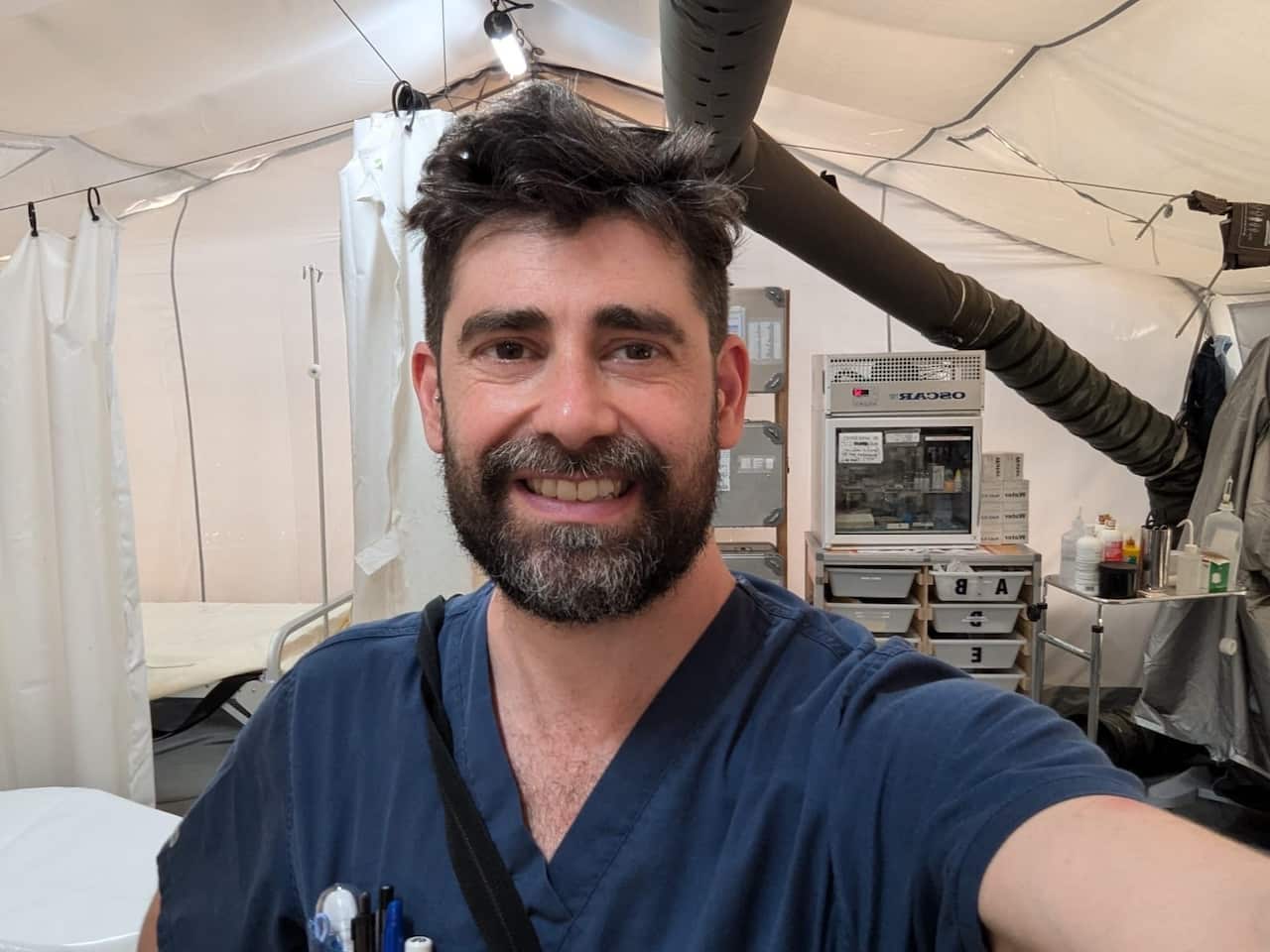 A bearded man in a blue nursing uniform smiles as he clicks a selfie inside a makeshift clinic.
