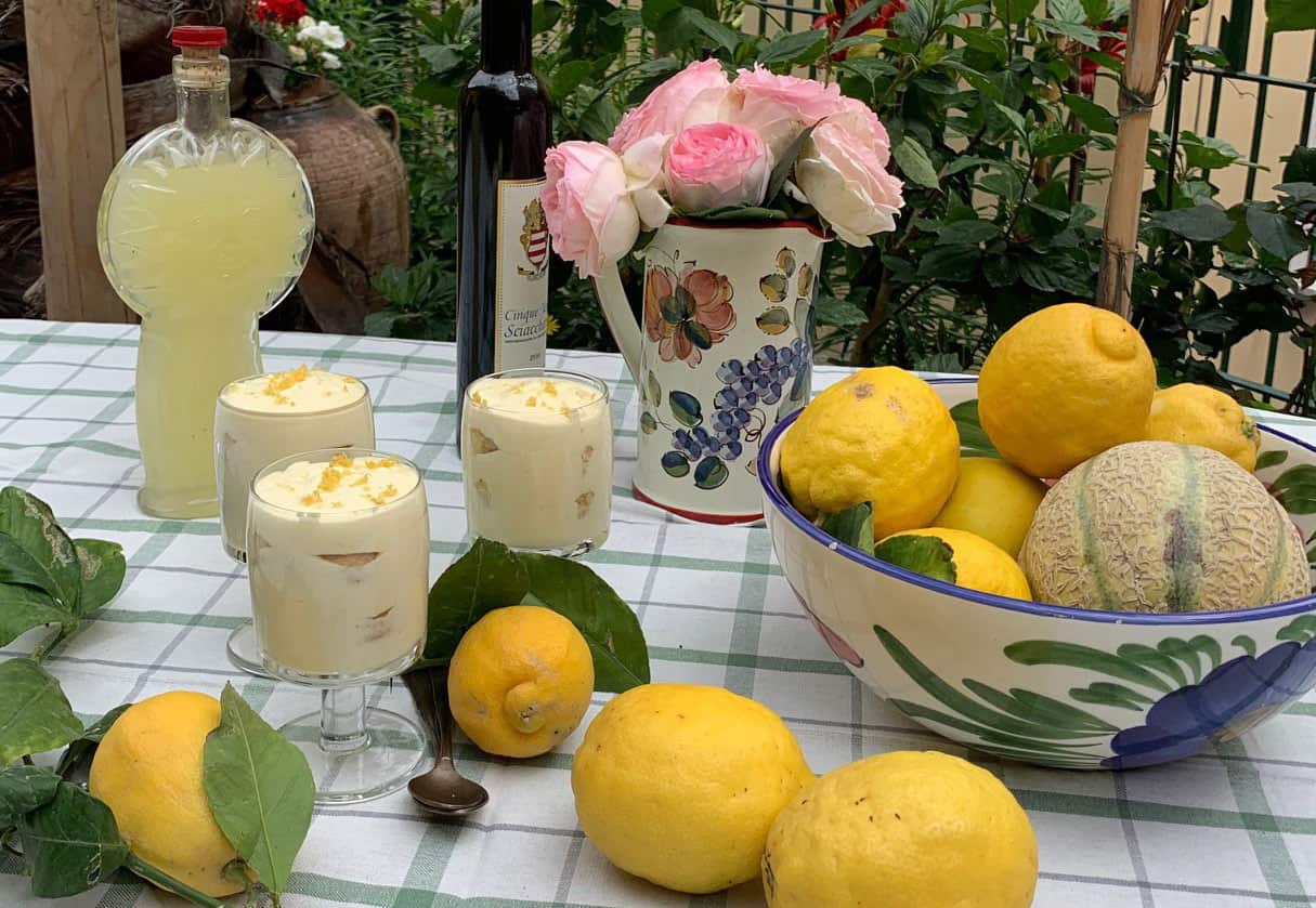 Three glasses filled with a creamy dessert sit on a table covered with a checked cloth. Lemons, a vase with flowers and a bottle of yellow liqueur also sit on the table.