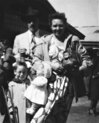 A man wearing a hat stands next to a woman in jacket and a child holding a doll.