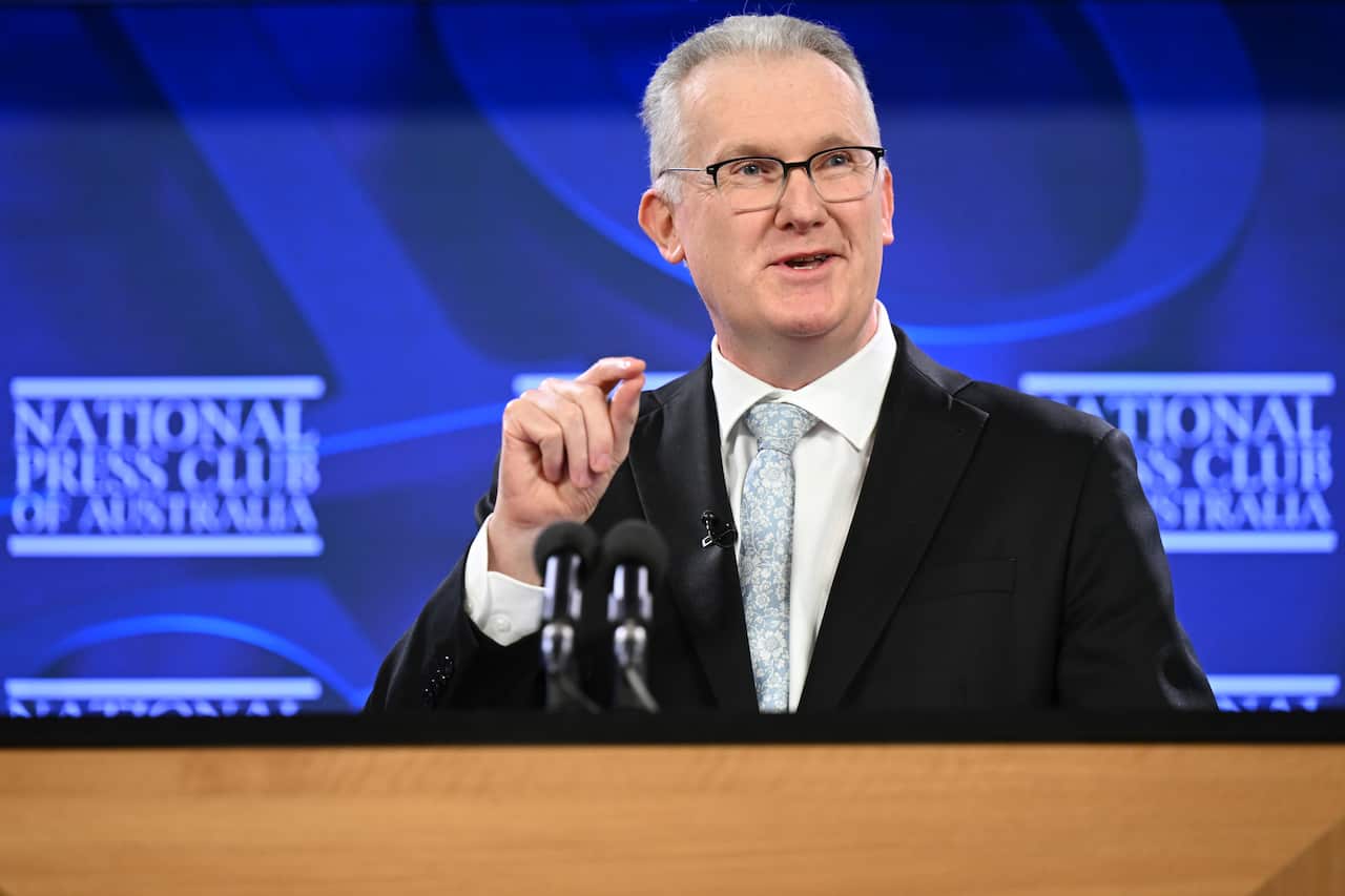 A middle-aged white man with grey hair and glasses, wearing a suit and tie. He is standing behind a microphone at a podium.