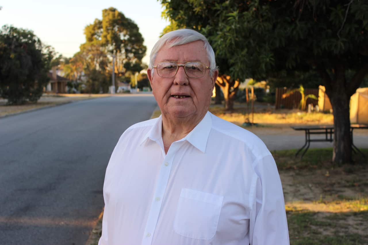 A man in a white shirt stands on a street with a serious expression on his face.