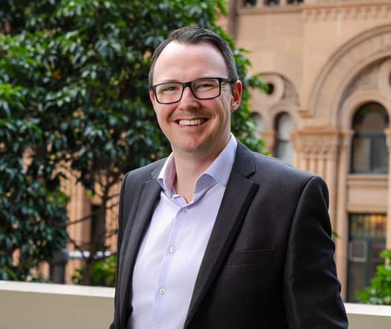A smiling man with short dark hair and black-rimmed glasses is wearing a dark grey suit jacket over a light grey button-down shirt, posed outdoors with lush green foliage and an ornate sandstone building blurred in the background.