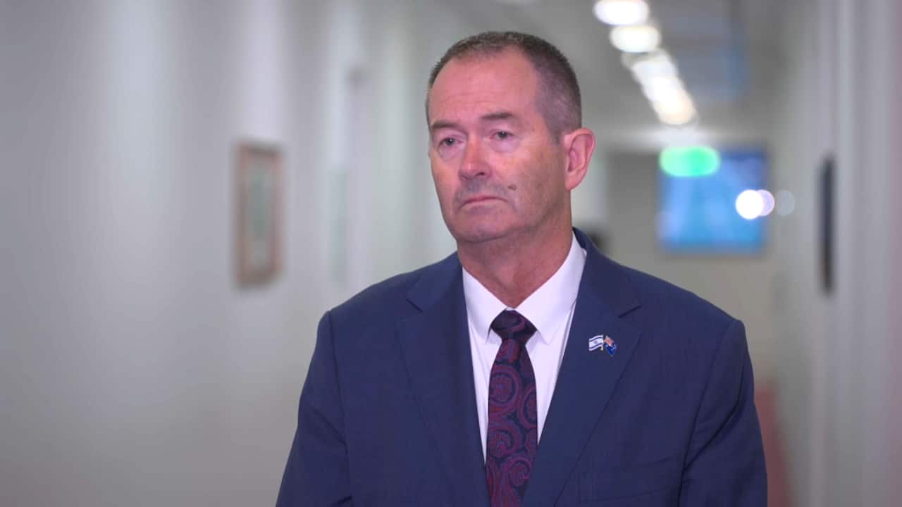 A man wearing a dark blue suit, standing in a hallway.