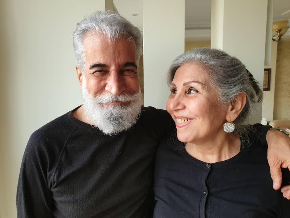 A man with a grey beard has his arm around a woman wearing a black top.