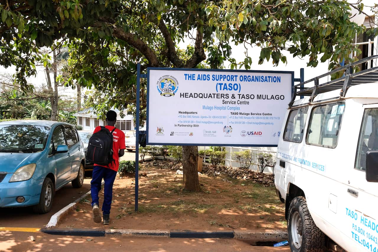 A man in a red shirt and a black backpack walks past an HIV clinic board with vehicles parked in the vicinity. 