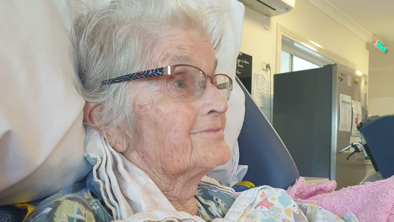 an older woman smiling looking into the distance, sitting rugged up in a hospital bed within an aged care centre
