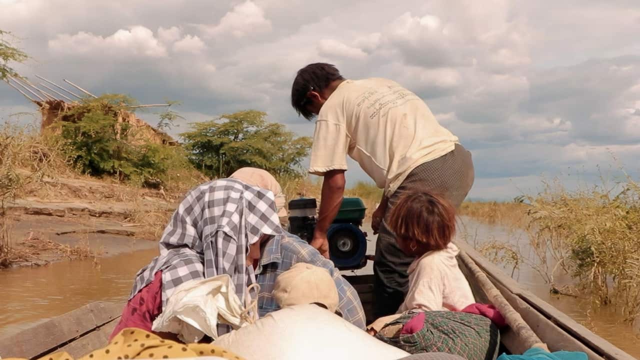 Several people travel in a narrow wooden boat along a muddy waterway bordered by dry vegetation. A man stands at the stern operating a small motor, while others sit among bags and belongings