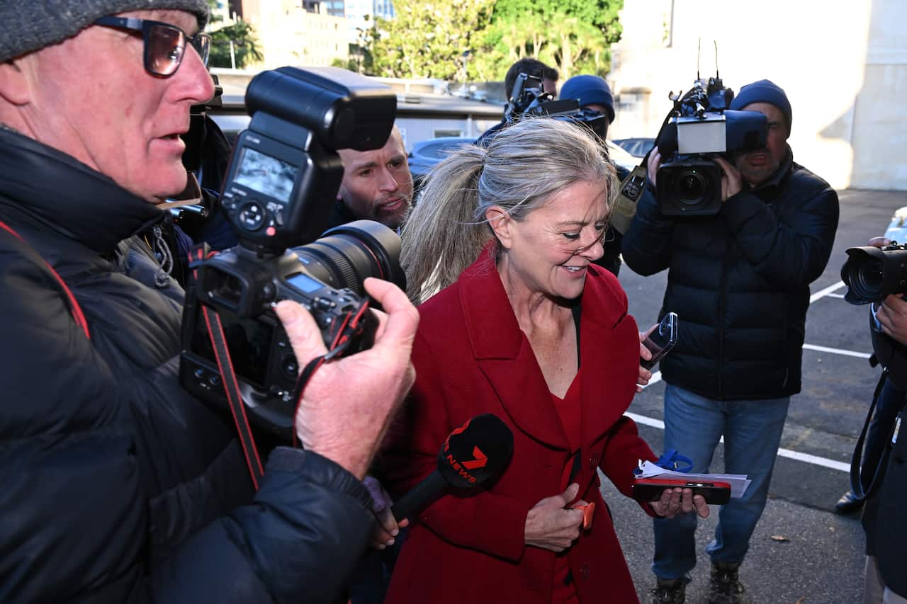 A woman in a bright red coat walks through a throng of journalists and photographers holding cameras and microphones toward her.