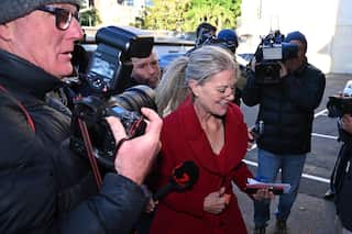 A woman in a bright red coat walks through a throng of journalists and photographers holding cameras and microphones toward her.