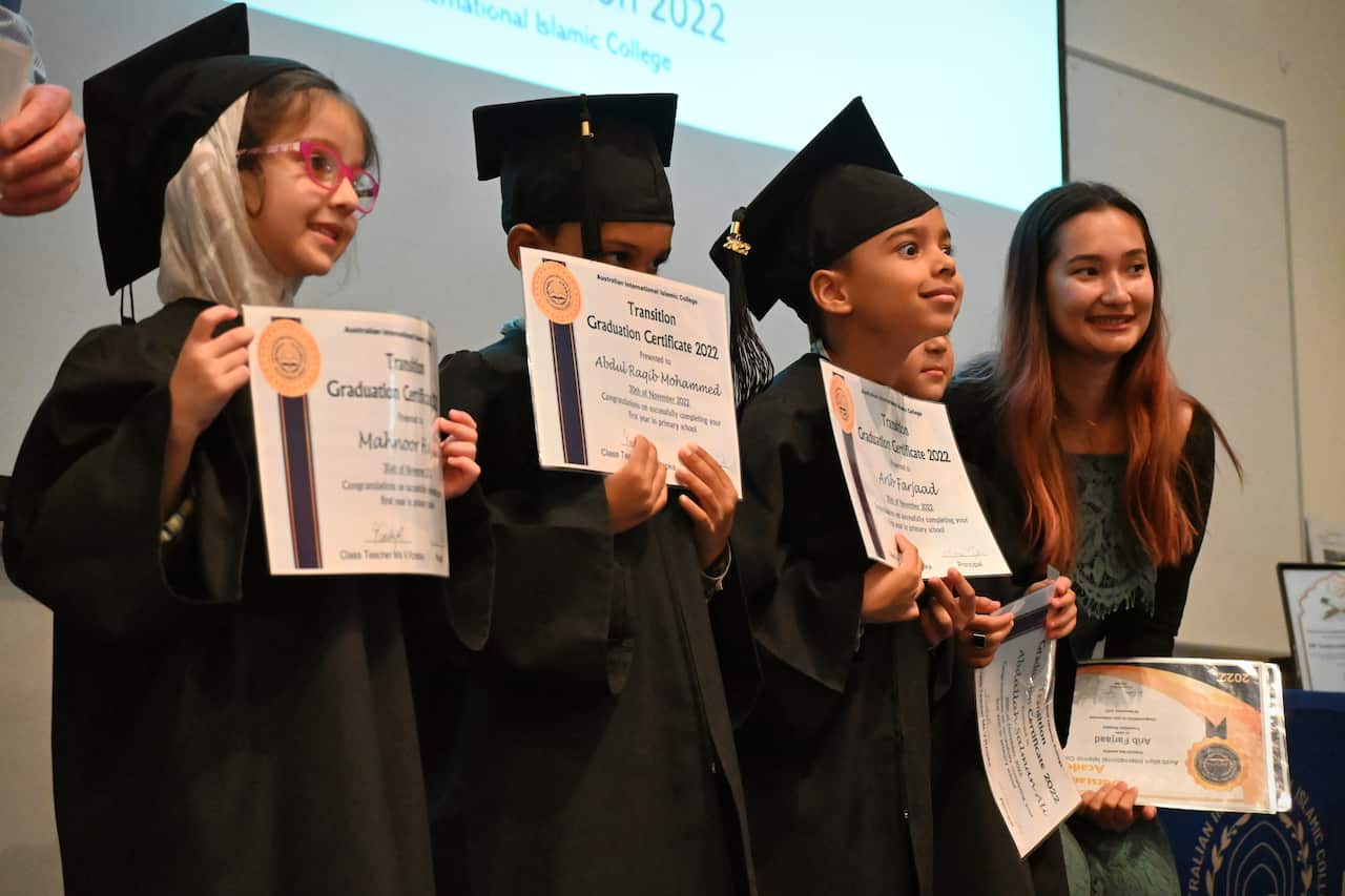 Three children wearing black academic gowns and mortar boards hold up certificates while standing next to a teacher holding a certificate.