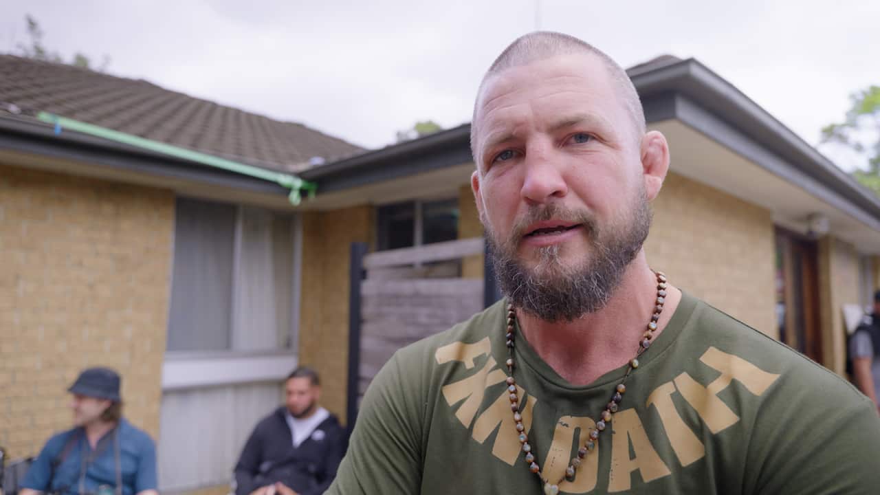 A man in a green t-shirt and wearing a cross on a beaded necklace looking down the camera in a backyard