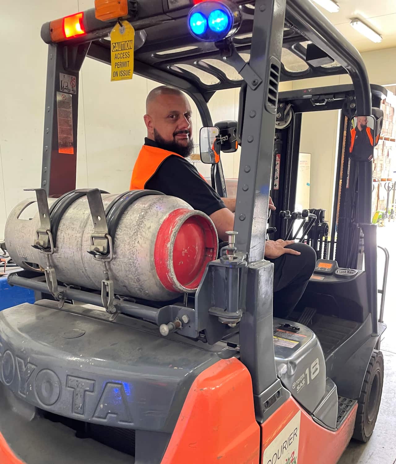 A man in a hi viz vest looking over his shoulder while sitting on a forklift at a warehouse.