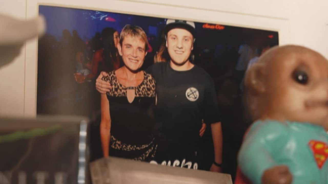 a photo in a frame on a bookshelf of a mother and a son in his early twenties. The son has his right arm around her shoulder and her left arm is around his waist