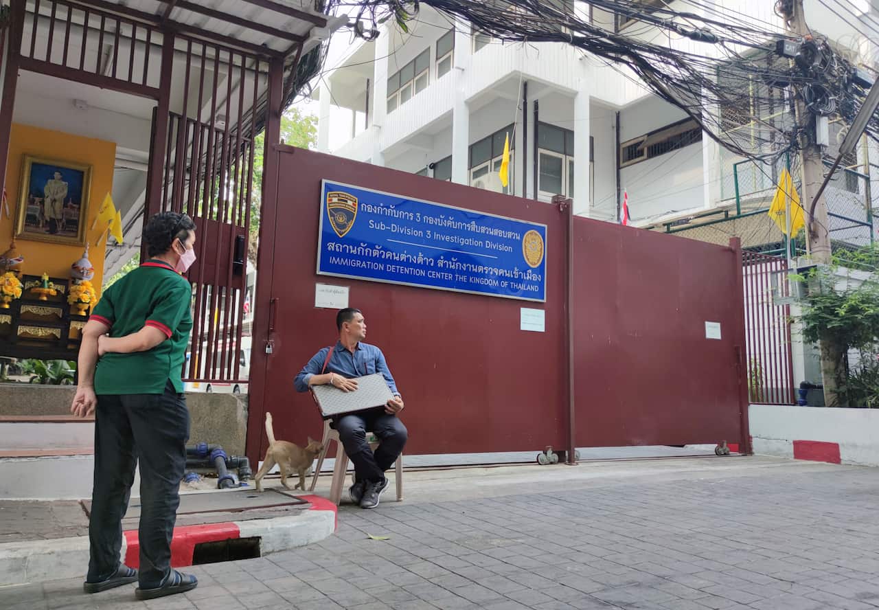 A gate with a blue sign on it. A man is sitting in a chair in front of it and another man is standing nearby