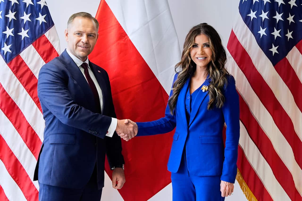 A man and a woman shaking hands in front of American and Polish flags.