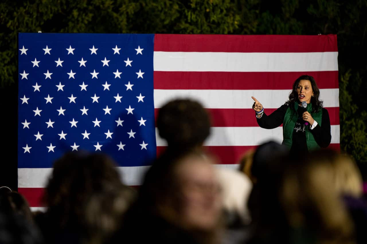 A woman in a green puffer vest talks into a microphone, in front of a large flag of the United States.