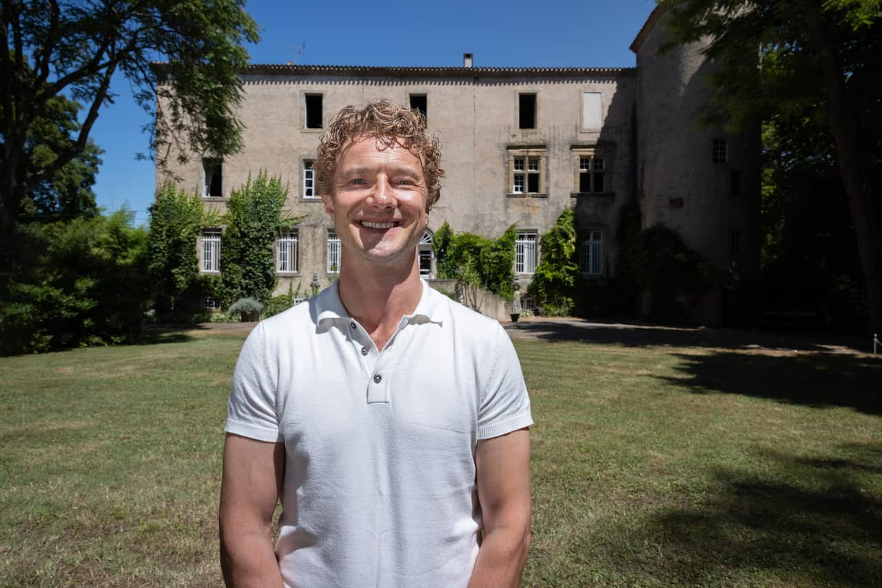 A smiling man with curly blond hair stands in front of a grand manor house.