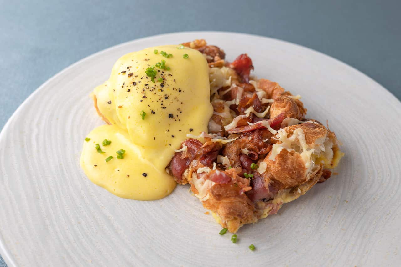 A rectangle of croissant and bacon 'pudding' sits on a plate. An egg covered in hollandaise sauce sits on top. 