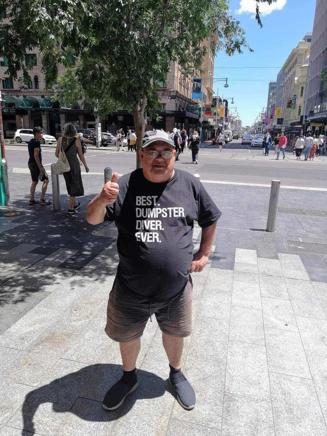 A man smiles in a hat and t shirt that reads "best dumpster diver ever" while holding a thumbs up on a busy street during the day