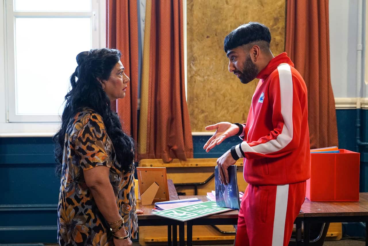A young man in a red tracksuit and a woman in a patterned dress face each other, apparently mid-conversation. 