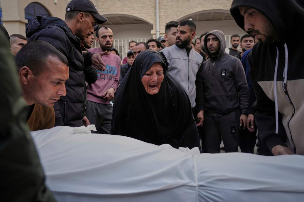 A woman cries over a body wrapped in a white sheet. There are people standing around her.