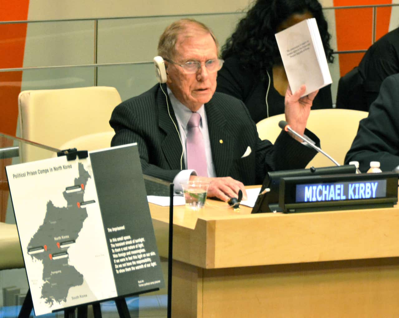 A man sitting at a court bench wearing headphones holds up a report. Next to him is a map of North Korea