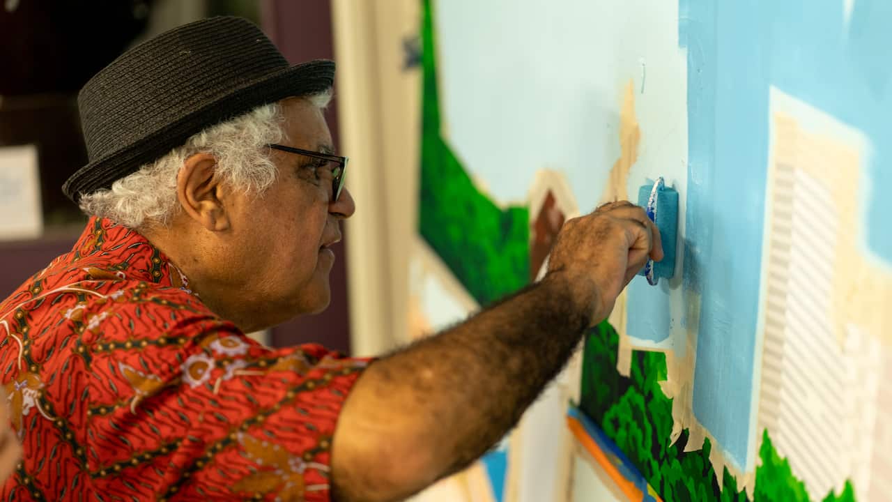 a close-up, side-on photo of richard bell, wearing a red shirt and black fedora, taking a roller of blue paint to a large canvass