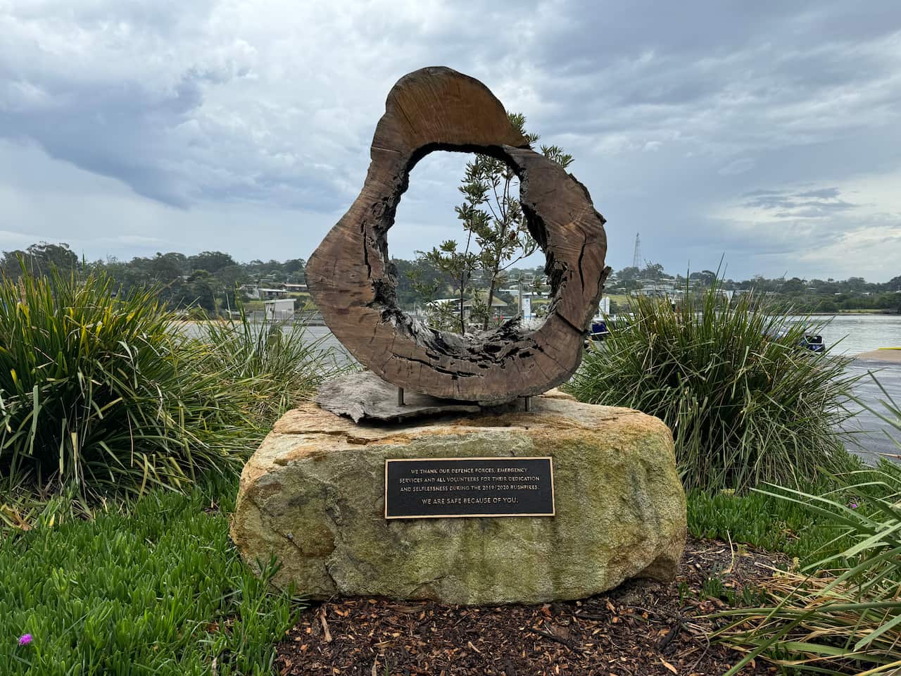 A log art memorial mounted atop a stone surrounded by bushes.